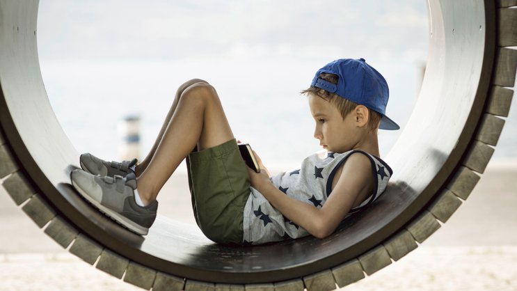 Ein kleiner Junge auf einem Spielplatz hat seinen Blick auf ein Smartphone vertieft.