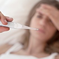Eine Frau hält sich die Stirn und betrachtet die Temperatur auf einem Fiebermesser.