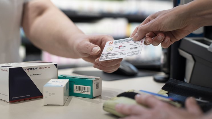 In der Apotheke: Eine Hand hält eine Sanitas-Krankenkasse, gegenüber die Hand der Kundin am Portemonnaie. Dazwischen liegen Medikamente auf dem Tisch.